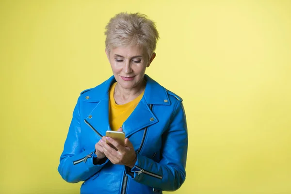 stylish woman at the age with a short hairstyle, in a blue jacket on a yellow background with a phone in her hands