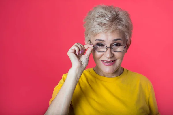 stylish aged woman with a short haircut and wearing glasses in a yellow T-shirt on a red background