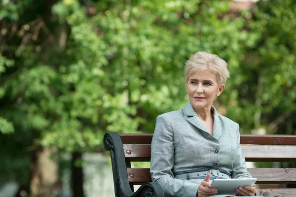 stylish elderly woman with a short haircut sitting on a bench with a tablet in hand in a park in summer