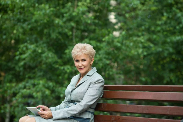 stylish elderly woman with a short haircut sitting on a bench with a tablet in hand in a park in summer