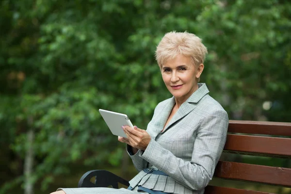 stylish elderly woman with a short haircut sitting on a bench with a tablet in hand in a park in summer