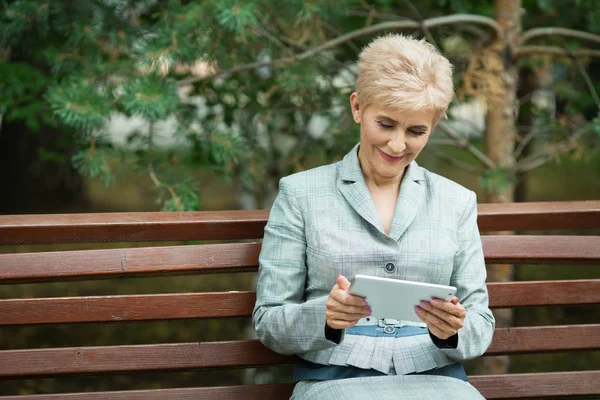 stylish elderly woman with a short haircut sitting on a bench with a tablet in hand in a park in summer