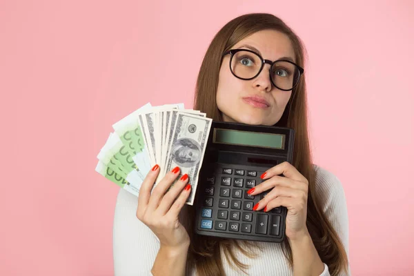 お金とピンクの背景の彼女の手で電卓をメガネで美しい少女 — ストック写真