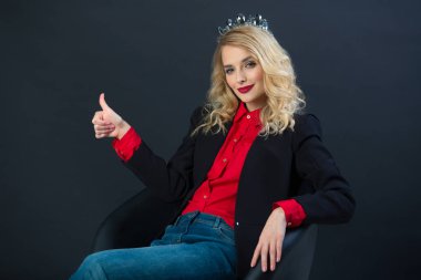 siyah ceketli güzel genç kız başını bir el hareketi ile siyah bir zemin üzerine bir taç ile bir sandalyede oturur