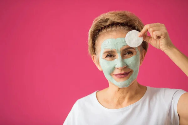 beautiful elderly woman with a wellness cream om face mask on a pink background with a cleansing napkin in hand