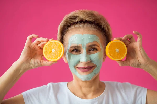 beautiful elderly woman with a wellness cream face mask on a pink background with an orange in her hand