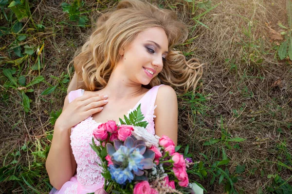 美丽的年轻女子化妆在粉红色的礼服躺在公园的草地上 一束花 — 图库照片