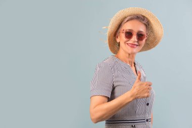 Güneş gözlüklü, mavi arka planda şapkalı, el hareketleriyle yaşlı bir kadın.