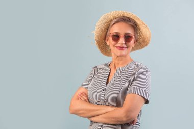 Güneş gözlüklü, mavi arka planda şapkalı yaşlı bir kadın.