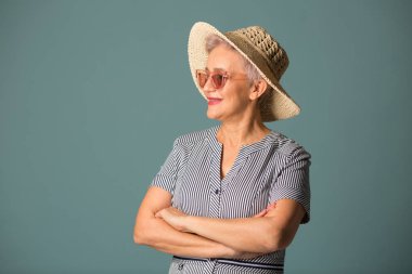 Güneş gözlüklü, mavi arka planda şapkalı yaşlı bir kadın.