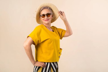 Güneş gözlüklü bej arka planda şapkalı yaşlı bir kadın.