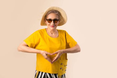 Güneş gözlüklü, bej arka planda şapkalı yaşlı bir kadın. El hareketleriyle.