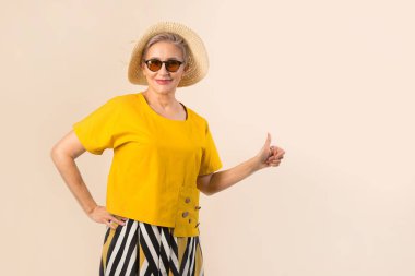 Güneş gözlüklü, bej arka planda şapkalı yaşlı bir kadın. El hareketleriyle.