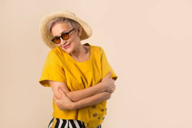 Güneş gözlüklü bej arka planda şapkalı yaşlı bir kadın.