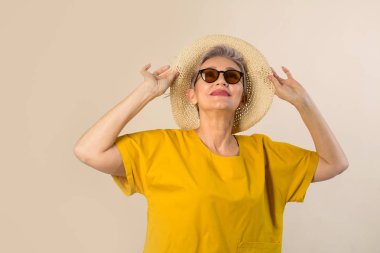 Güneş gözlüklü bej arka planda şapkalı yaşlı bir kadın.