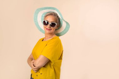 Güneş gözlüklü bej arka planda şapkalı yaşlı bir kadın.