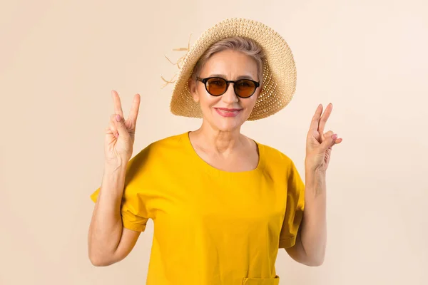 adult old woman in a hat on a beige background in sunglasses with hands up