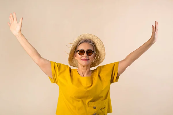 adult old woman in a hat on a beige background in sunglasses with hands up
