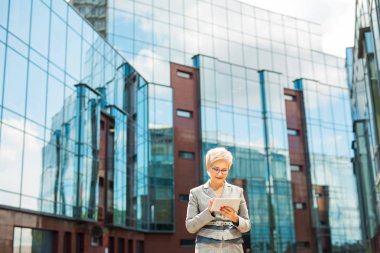 Takım elbiseli ve gözlüklü şık bir yetişkin kadın elinde tabletle cam bir binanın arka planına yaslanmış.