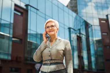  Takım elbiseli ve gözlüklü şık yetişkin bir kadın cam bir binanın arka planında bir telefon görüşmesi yapar.