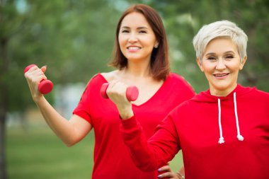 İki güzel yetişkin kadın yaz parkında dambıllarla spor yapıyor.