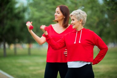 İki güzel yetişkin kadın yaz parkında dambıllarla spor yapıyor.