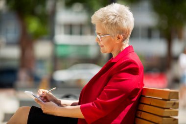 Kırmızı ceketli güzel, şık bir kadın bir bankta oturur ve bir deftere yazar.