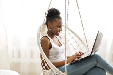  Güzel, genç bir Afrikalı kadın asılmış bir sandalyede dizüstü bilgisayarla oturuyor.