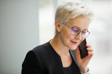 Yaşı ilerlemiş güzel, şık bir kadın. Siyah takım elbiseli, telefonla konuşuyor.