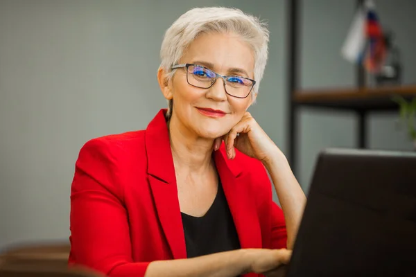 Kırmızı ceketli, şık, yaşlı bir kadın ofiste dizüstü bilgisayarla çalışıyor.