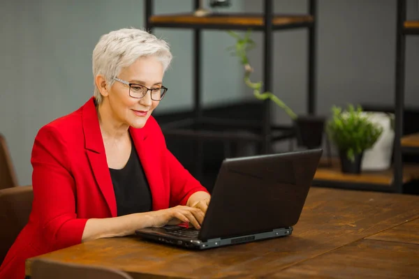 Kırmızı ceketli, şık, yaşlı bir kadın ofiste dizüstü bilgisayarla çalışıyor.