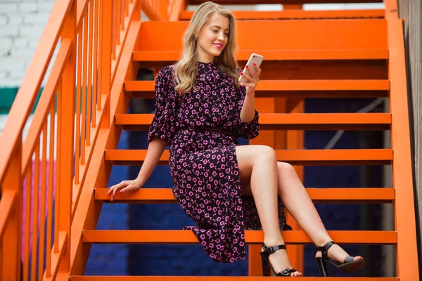 漂亮的年轻女人穿着裙子站在明亮的橙色楼梯旁边 带着电话 — 图库照片