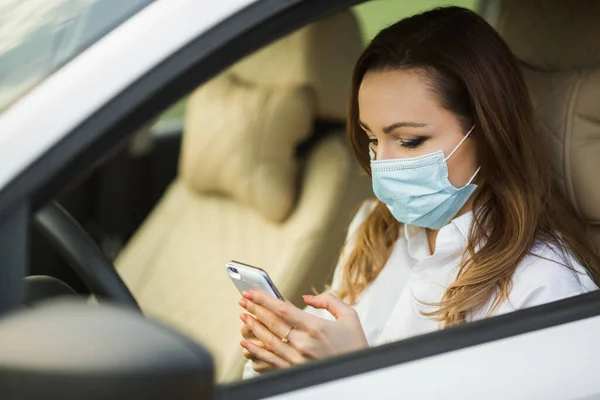 beautiful young woman in a medical mask driving a car with a mobile phone