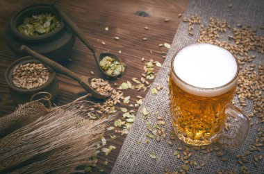 Soğuk köpük bira malt tahıl ile yeşil hop tencere ve çavdar kulaklar kahverengi ahşap masa arka plan üzerine çuval bezi kumaş yaprak.