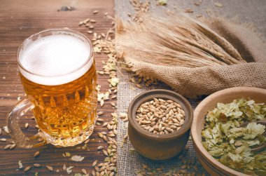 Soğuk köpük bira malt tahıl ile yeşil hop tencere ve çavdar kulaklar kahverengi ahşap masa arka plan üzerine çuval bezi kumaş yaprak.
