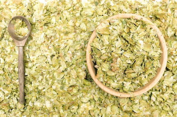 Granulated green hop leaves in the wooden bowl with wood spoon on scattered hop leaves background.