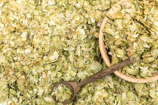 Granulated green hop leaves in the wooden bowl with wood spoon on scattered hop leaves background.