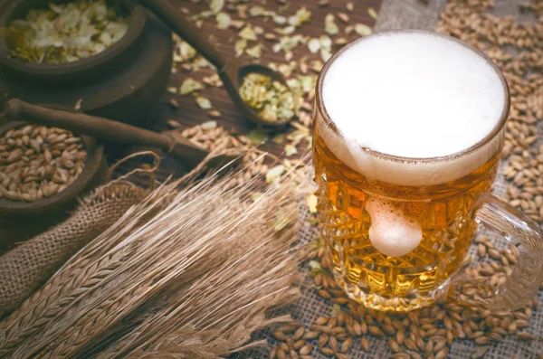 冷たい泡ビール麦芽穀物 緑ホップの葉ポットやライの耳に茶色の木製のテーブル背景に黄麻布の上 — ストック写真