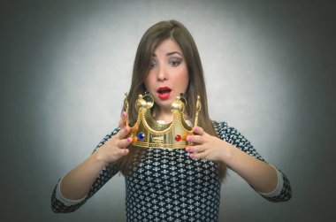 Sürpriz ve heyecanlı bir kadın kız altın taç Ödülü gri arka plan üzerinde izole elinde tutuyor. Kazanan. İlk yer kavramı.