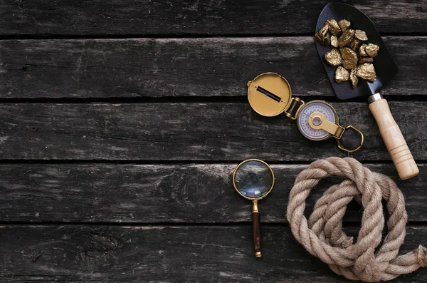 Shovel full of gold ore, compass, rope and magnifying glass on aged wooden table background. Treasure hunter concept.
