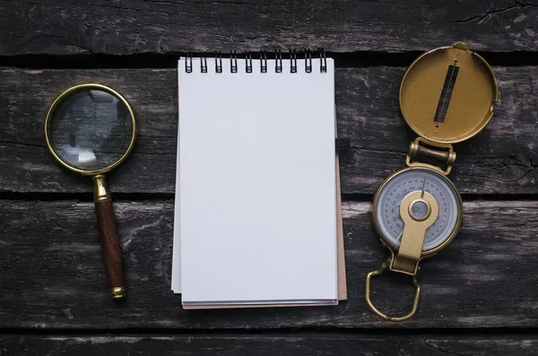 Magnifying glass, compass and blank page notepad on old aged wooden table background.