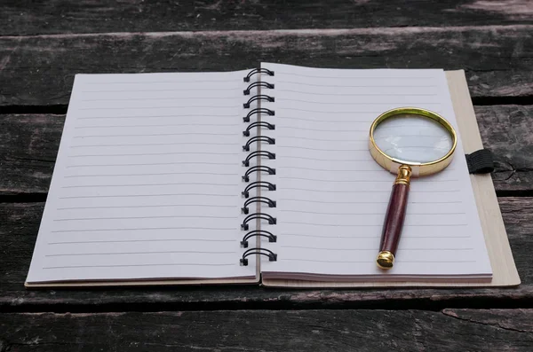 Magnifying glass and blank page notepad on old aged wooden table background.