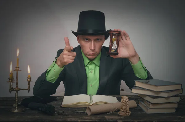 Severe detective agent man in a bowler hat is sitting by the table and holding in hands a sand clock. Detective or spy agent or police inspector or mafia boss concept.