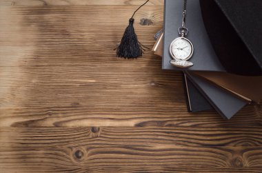 Lisansüstü kap, cep saati ve kopya alanı ile ahşap okul masasında kitap yığını.