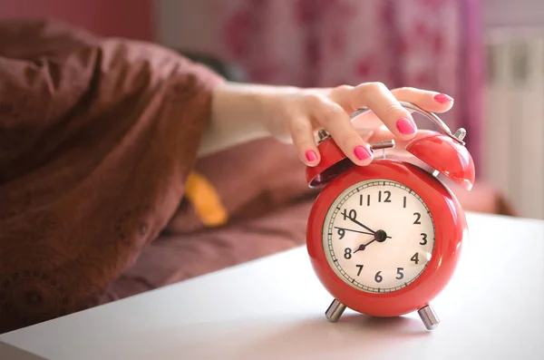 Sleepy woman in the bed is trying to turn off a red alarm clock on the table at morning time.