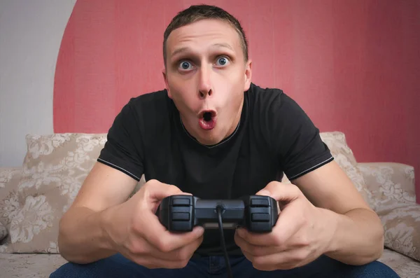 Surprised and enthusiastic gamer man with a joystick in his hands is sitting on a sofa with wide open mouth and is playing in video game. Exciting game concept