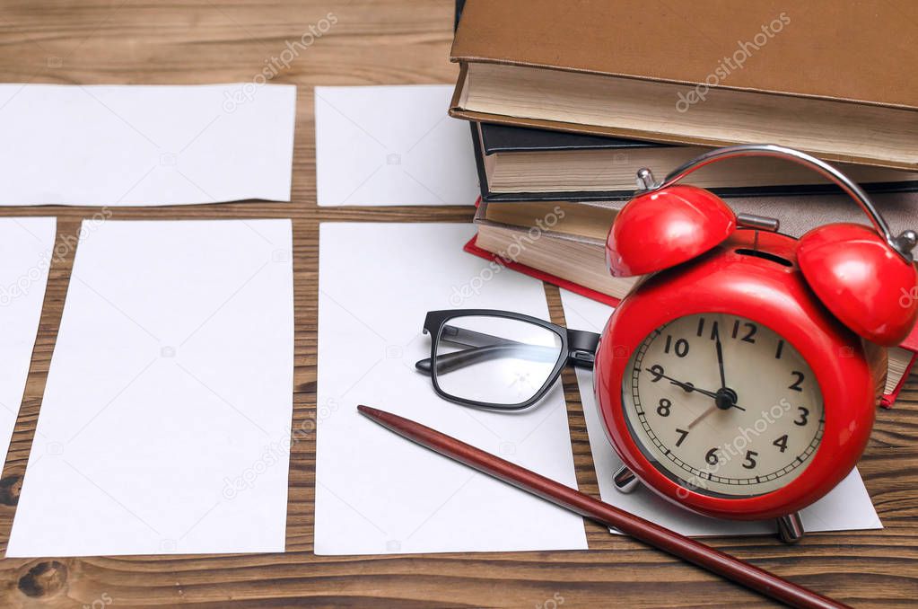 Blank exam tickets, books, school pointer, glasses and a red alarm clock on the school desk in the classroom. Education background. Time to test work.