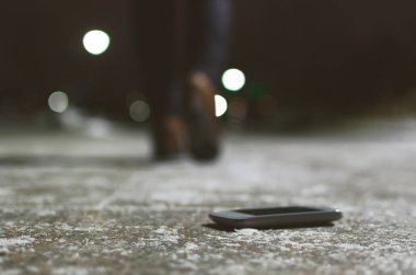 Lost mobile phone dropped from a woman pocket on the night footpath and is walking away silhouette of woman. clipart