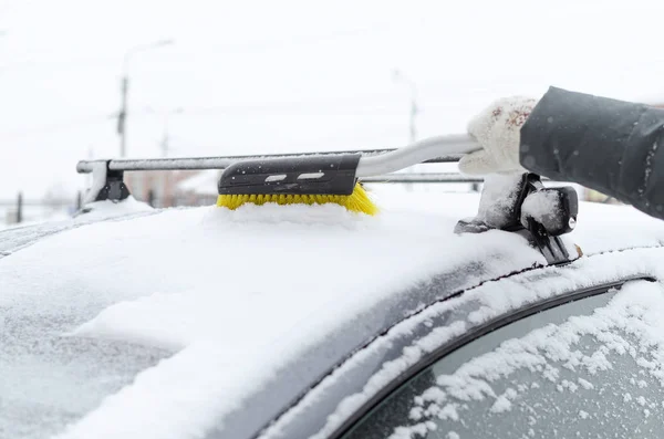 The driver cleans the car from the snow with a brush on the winter street.