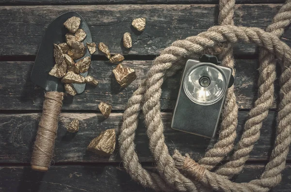 Gold nuggets on the spade, rope and flashlight on the aged wooden table background. Treasure hunter or goldminer concept.